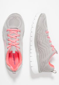 Hellgraue Sportschuhe mit pinken Akzenten, strukturiertem Netzobermaterial, Schnürverschluss und einer weißen, gepolsterten Sohle mit Zickzackmuster.