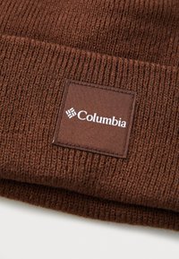 Ruda megzta kepurė su tekstūruotu paviršiumi, kurioje yra kvadratinė medžiagos etiketė su balta "Columbia" ženklų.
