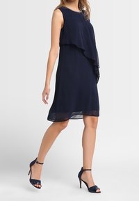 Nawigacyjna sukienka z szyfonu o warstwowej konstrukcji; bez rękawów z zaokrąglonym dekoltem. W połączeniu z czarnymi sandałami na wysokim obcasie i zwiewnym dołem.