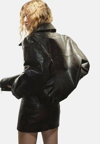 Chaqueta bomber de piel negra con un ajuste ligeramente holgado, costuras visibles y un cuello alto. Textura suave y acabado brillante.