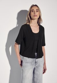 Cardigan nero a maniche corte con una vestibilità ampia e scollatura a V, indossato sopra una canotta nera. Abbinato a jeans in denim chiaro.