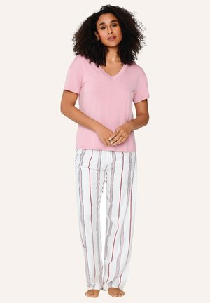 Femme aux cheveux bouclés portant un T-shirt rose clair à col en V et un pantalon blanc avec des rayures verticales rouges et grises, debout pieds nus.