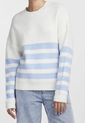 Femme portant un pull en maille blanc avec des rayures bleu clair sur la poitrine et les bras, assorti à un jean de couleur claire.