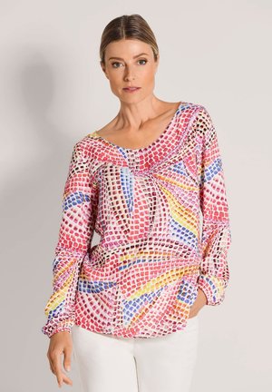 Frau trägt langärmeliges, mehrfarbiges, geometrisch gemustertes Oberteil mit gerafftem Ausschnitt, kombiniert mit weißen Hosen, steht vor einfachem Hintergrund.