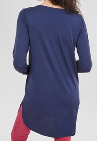 Tunique bleu marine à manches longues avec un col rond et des fentes latérales. Le tissu semble doux et extensible. Portée par-dessus un pantalon rouge.