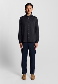 Camicia nera a maniche lunghe con un motivo sottile, bottoni frontali, colletto a punta e un piccolo logo. Abbinata a pantaloni blu navy e scarpe beige.