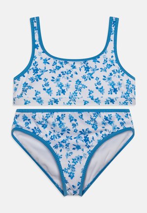 Dwuczęściowy strój kąpielowy z białym tłem i niebieskimi kwiatowymi wzorami, niebieskimi wykończeniami i gładką teksturą. Górna część ma szerokie ramiączka, a dolna jest wycięta wysoko.