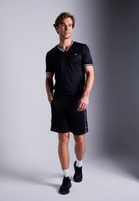 Svart sportoutfit med kortärmad V-ringad tröja med vit kant och matchande shorts. Bärs med vita strumpor och svarta sneakers.