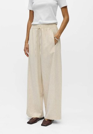 Kobieta ubrana w luźne beżowe lniane spodnie ze ściągaczem w talii, biały krótki rękaw, oraz ciemnobrązowe buty z kwadratowym noskiem.
