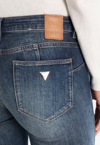 Jeans de mezclilla azul oscuro con una etiqueta de cuero marrón que presenta texto en relieve. El bolsillo trasero tiene un acento de logo en forma de triángulo plateado. Diseño clásico de cinco bolsillos.