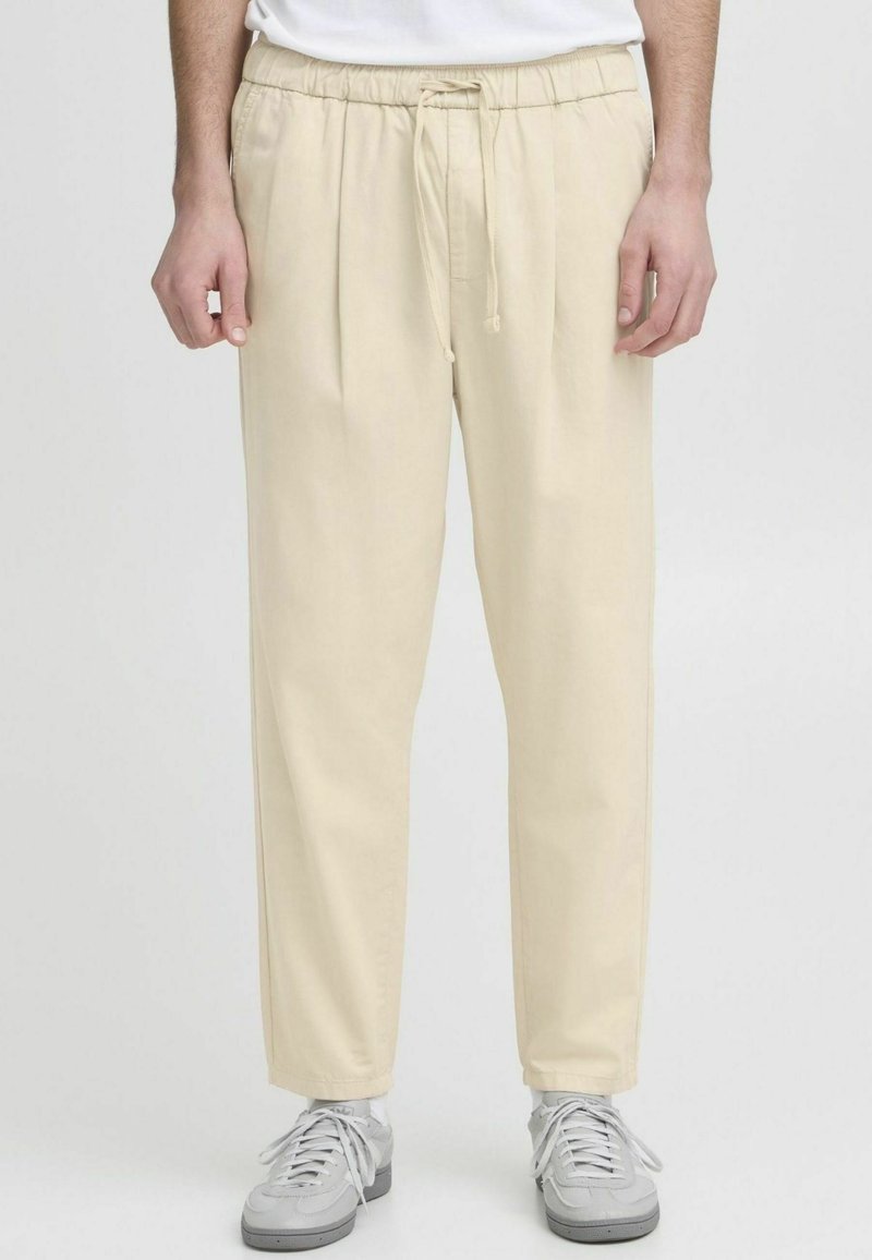 Homme portant un pantalon beige à cordon de serrage et des baskets blanches, debout avec les mains détendues le long du corps sur un fond neutre.