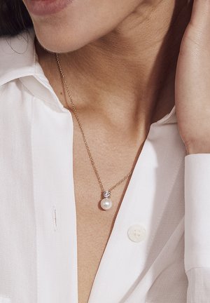Goldkette mit einer runden Perlenanhänger, akzentuiert mit einem kleinen klaren Edelstein, getragen über einem weißen Hemd mit Kragen.