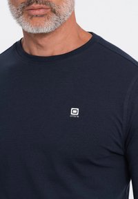 Tummansininen pitkähihaainen paita sileästä kankaasta, jossa on pieni valkoinen logo rinnassa. Klassinen pyöreä pääntie, jossa on ribbireuna.