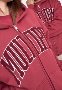 Sudadera con cremallera en color burdeos que presenta grandes letras en blanco y burdeos texturizadas, puños acanalados y capucha con cordón. Tejido suave con un acabado liso.