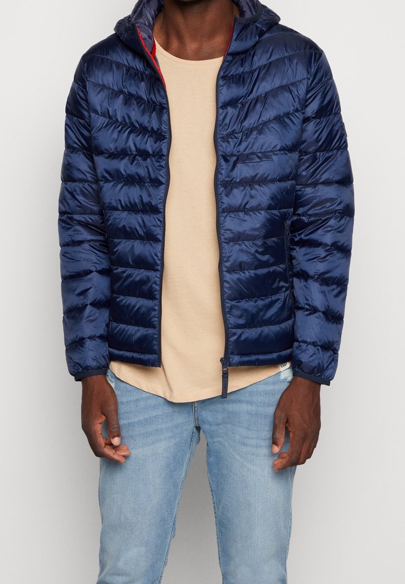 Giubbotto puffer blu navy con una texture liscia e trapuntata, dotato di cappuccio, con dettagli rossi lungo la cerniera e una vestibilità slim.