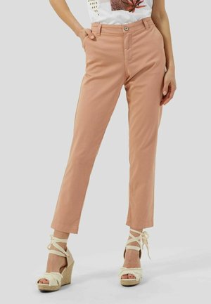 Kobieta ubrana w jasnoróżowe spodnie o prostych nogawkach i białe sandały na koturnie z paskami wokół kostek, stojąca z częściowo wsuniętymi rękami do kieszeni.