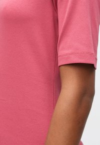 Δεξί χέρι ατόμου που φορά ροζ μπλουζάκι κοντομάνικo με ορατές ραφές στο στρίφωμα των μανικιών, απέναντι από λευκό φόντο.