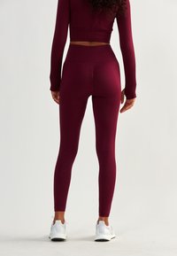 Bordowe legginsy z wysokim stanem, o teksturowanym wzorze, oraz białe buty sportowe, widziane od tyłu na tle w neutralnym kolorze.