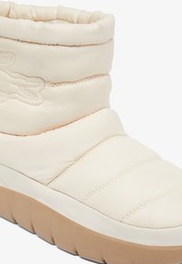 Botín acolchado de color crema con parte superior de tela texturizada, que presenta detalles de costura y una suela de goma en color marrón. Incluye un sutil acento de logo.