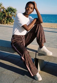 Junge Person sitzt auf einer Betonbank am Strand, trägt weiße Sneakers, eine Leoparden-Print-Hose mit pinken Streifen und ein weißes T-Shirt.