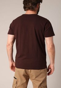 Camiseta de algodón marrón con mangas cortas, escote redondo y un corte relajado, vista desde la parte posterior, combinada con pantalones cargo caqui.