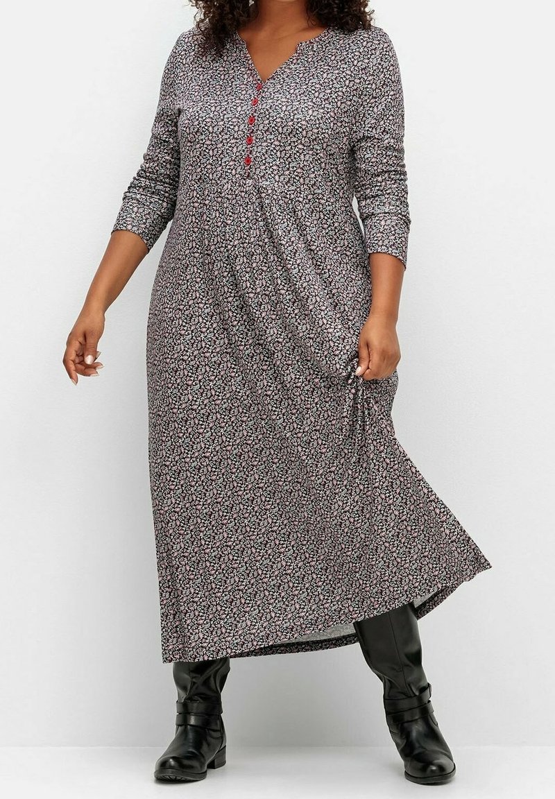 Femme portant une longue robe fleurie avec des boutons rouges et des bottes noires montant jusqu'aux genoux sur un fond blanc uni.