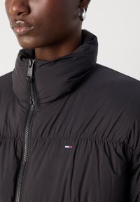 Svart lett dunjakke med høy krage, glidelås foran og samlet stoffdetaljer. Har en liten logo i rødt, hvitt og blått.