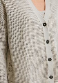 Light beige knitted cardigan with a V-neck, featuring a loose, open weave texture, five black buttons, and ribbed cuffs and hem.