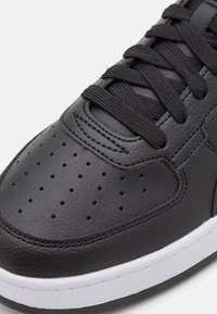 Černé kožené tenisky s hladkým povrchem, perforovanou špičkou, plochými černými tkaničkami a bílou gumovou podrážkou. Klasický nízký design.