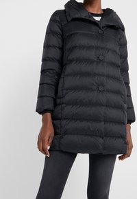 Sort quiltet pufferjakke med store knapper, båret over mørke jeans, der viser længde til midt på låret og lange ærmer.
