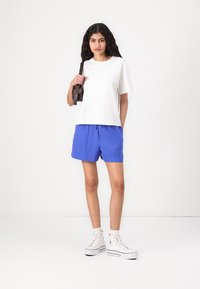 Hvit kortermet T-skjorte kombinert med blå shorts. Modellen har på seg hvite høye sneakers og holder en liten mørk håndveske.