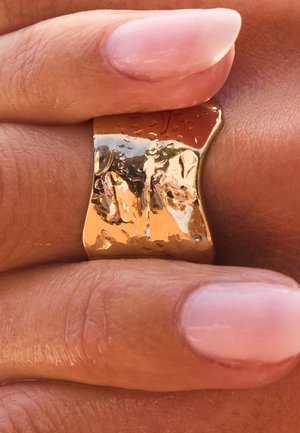 Breiter, gehämmerter Goldring an einem Finger mit glatter, natürlich getönter Haut und sorgfältig manikürten Nägeln mit blassrosa Nagellack.