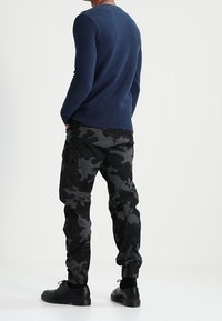 Marineblauer Strickpullover mit Rippstruktur kombiniert mit schwarzen Jogginghosen im Camouflage-Stil, die Cargotaschen und elastische Bündchen haben. Schwarze Schuhe runden den Look ab.