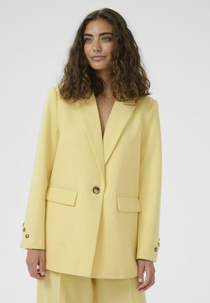 Ženska s kodrastimi rjavimi lasmi v pastelno rumenem suknjiču z gumbi in ujemajočih hlačah, oblečena na enostavni svetli podlagi.