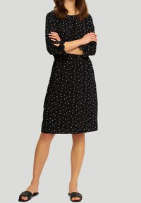 Robe noire à pois blancs, manches 3/4 avec liens, taille élastique, longueur genoux, assortie avec des sandales noires à enfiler.