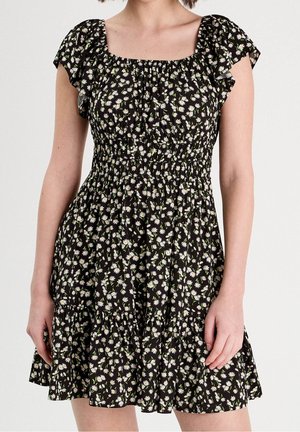 Femme portant une courte robe noire avec un motif floral de marguerites blanches et jaunes et des manches courtes volantées, debout devant un fond uni.