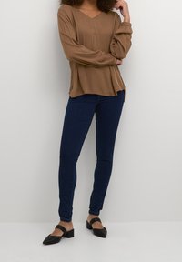 Bruine lange mouw top met een V-hals en een losse pasvorm, gecombineerd met donkerblauwe skinny jeans en zwarte blokhak schoenen.