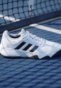Biela športová obuv Adidas s čiernymi pásmi na modrom tenisovom kurte pri sieti vrhajúca tieň na zem.