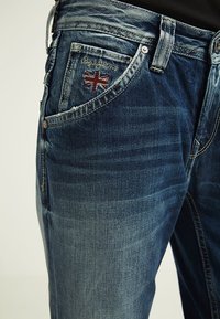 Denimjeans i mörkblått med blekta områden. Har en broderad Union Jack på framfickan och en klassisk femficksdesign.