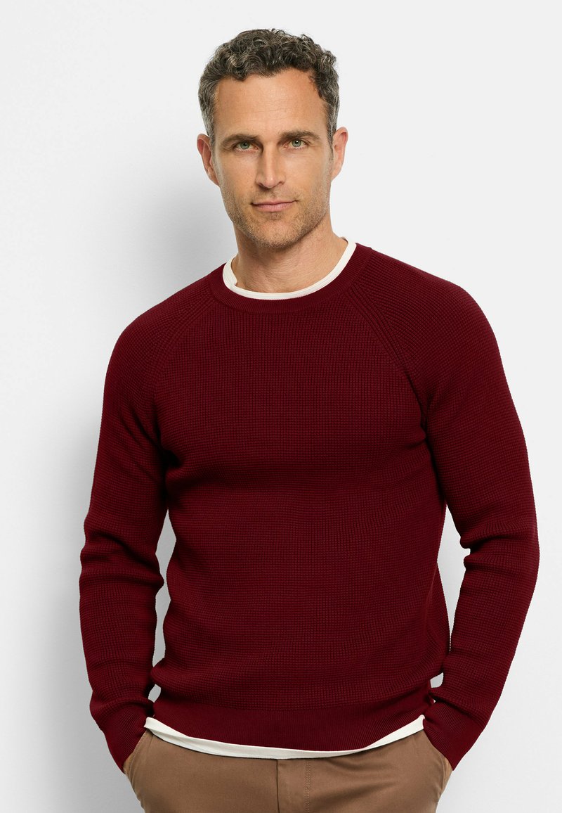 Burgundy knit sweater with a textured pattern, round neckline, and ribbed cuffs. Worn over a white shirt, features a fitted silhouette.