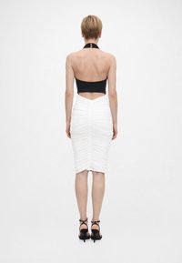 Γυναίκα που στέκεται πλάτη στην κάμερα, φορώντας μαύρο κορυφαίο halter, λευκή φούστα μέσης με ρυτιδώσεις και μαύρα ψηλοτάκουνα σανδάλια, σε απλό λευκό φόντο.