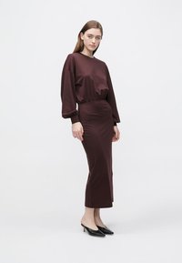 Bordó šaty s dlouhými rukávy, vyznačeným pasem a rozpínací sukní. Doplňují je černé lodičky s špičatou špičkou. Hladká textura, minimální design.