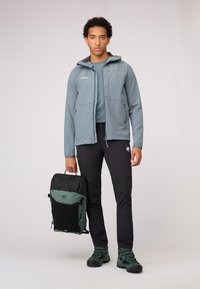 Grijze softshell jas met een capuchon, zwarte broek en groene wandelschoenen. Houdt een zwarte en groene rugzak met meerdere vakken vast.