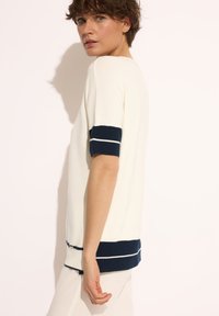 Kremowy sweterek dzianinowy z granatowymi paskami na mankietach i u dołu. Krótkie rękawy, luźny krój, gładka faktura. Zaprojektowany do noszenia w codziennych stylizacjach.
