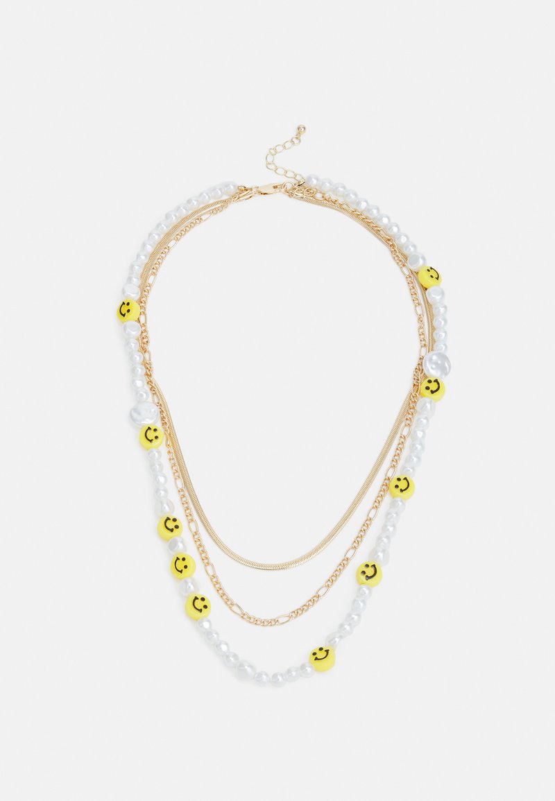 Colar multi-camadas em tom dourado, com pérolas brancas e contas amarelas em forma de rosto sorridente, destacando uma mistura de texturas e elementos de design divertidos.