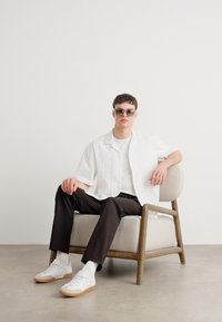 Witte kortekorte mouw shirt, zwarte broek, witte sneakers met tan kleurige zolen. Duidelijke bril. Zittend op een beige gestoffeerde stoel met houten poten.