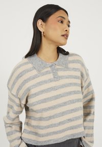 Młoda kobieta w swetrze w beżowo-szare paski z kołnierzykiem i guzikami, nosząca małe kolczyki w kształcie kół, patrząca w bok na tle jednolitego tła.