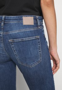 Mörkblå denimjeans med en slät textur, med en rektangulär läderlapp vid midjan och två bakfickor med vinklade kanter.