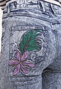 Dżinsy denimowe w wyblakłym niebieskim kolorze z wyraźnym haftem przedstawiającym fioletowy kwiat i zielone liście na tylnej kieszeni.