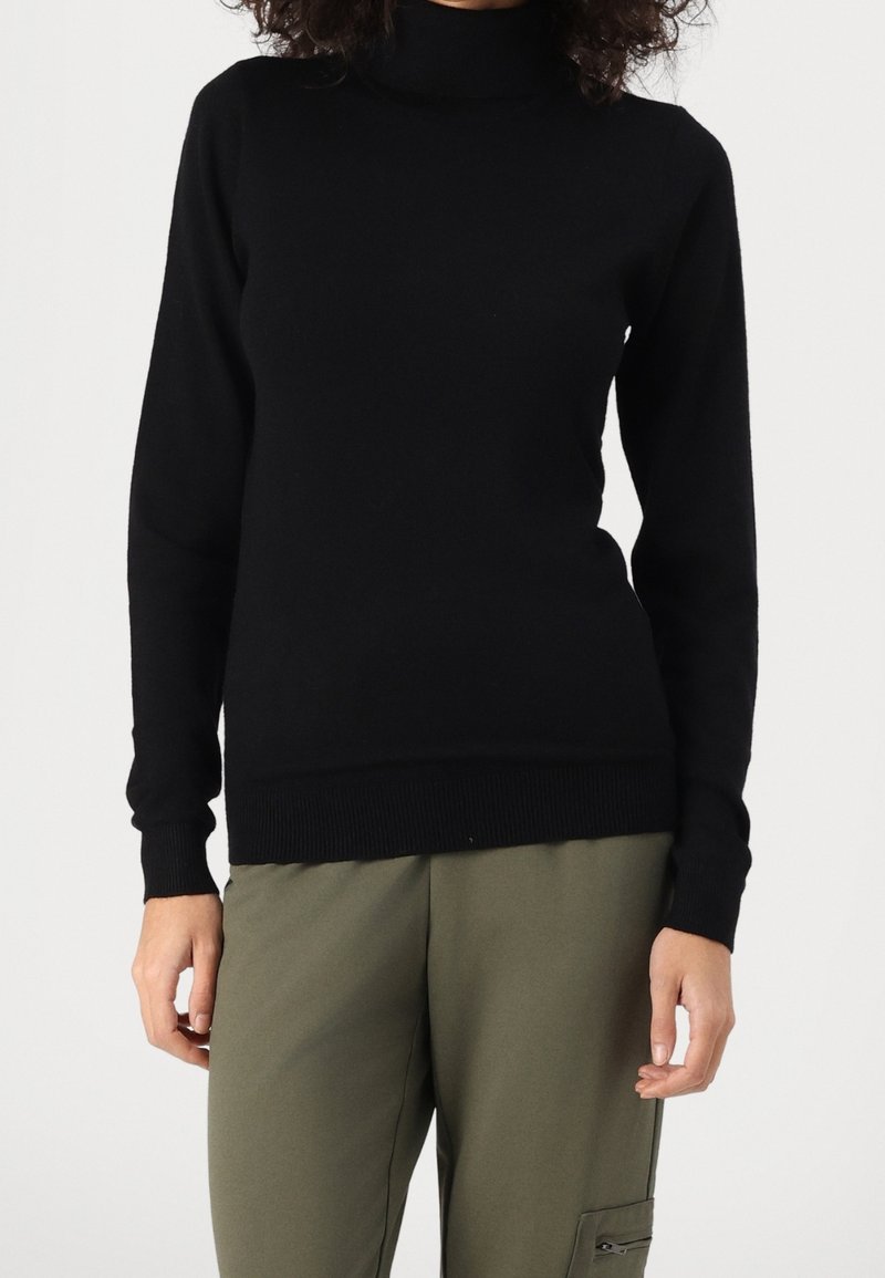 Personne portant un pull noir à col roulé et un pantalon vert olive avec une poche zippée sur le côté, debout devant un fond uni.